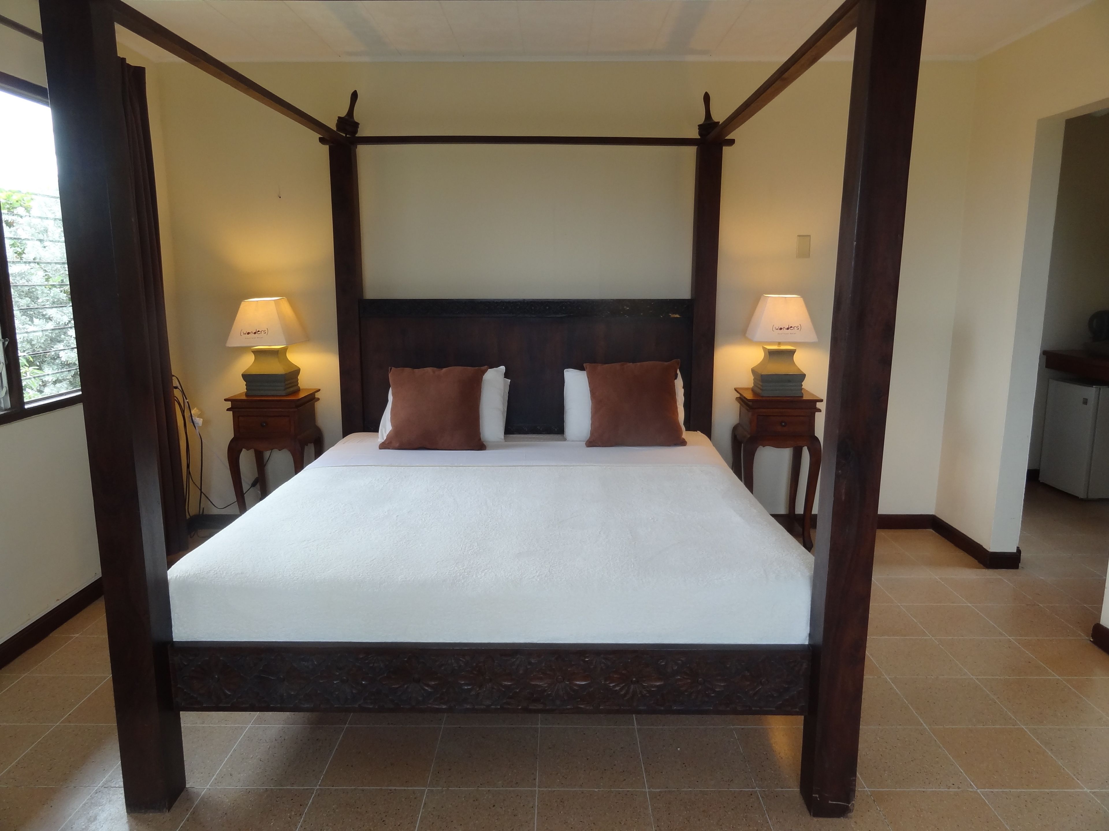 A four-poster bed with a dark wooden frame and white bedding, flanked by two bedside tables with lamps, in a room with tiled flooring.