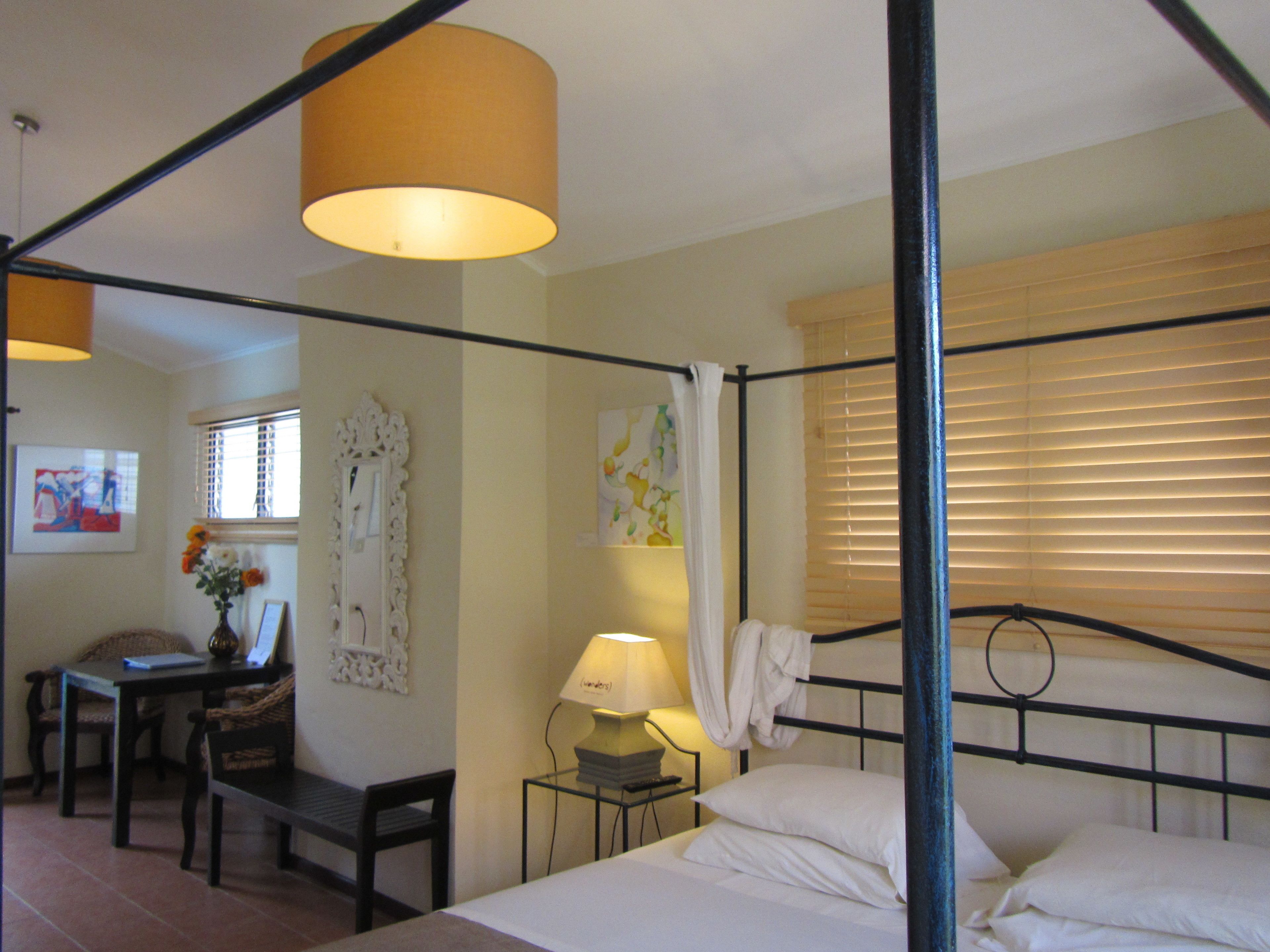A four-poster bed with white linens, a small bedside table with a lamp, a chair, and a desk with a vase of flowers.