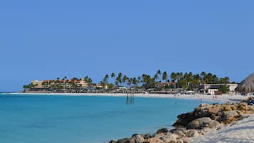 Un complejo turístico en la playa con orilla arenosa, agua azul cristalina y una hilera de edificios con palmeras.