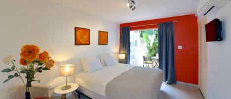 Habitación de hotel con una cama, una mesita con un florero, una silla, una lámpara y vistas al exterior.