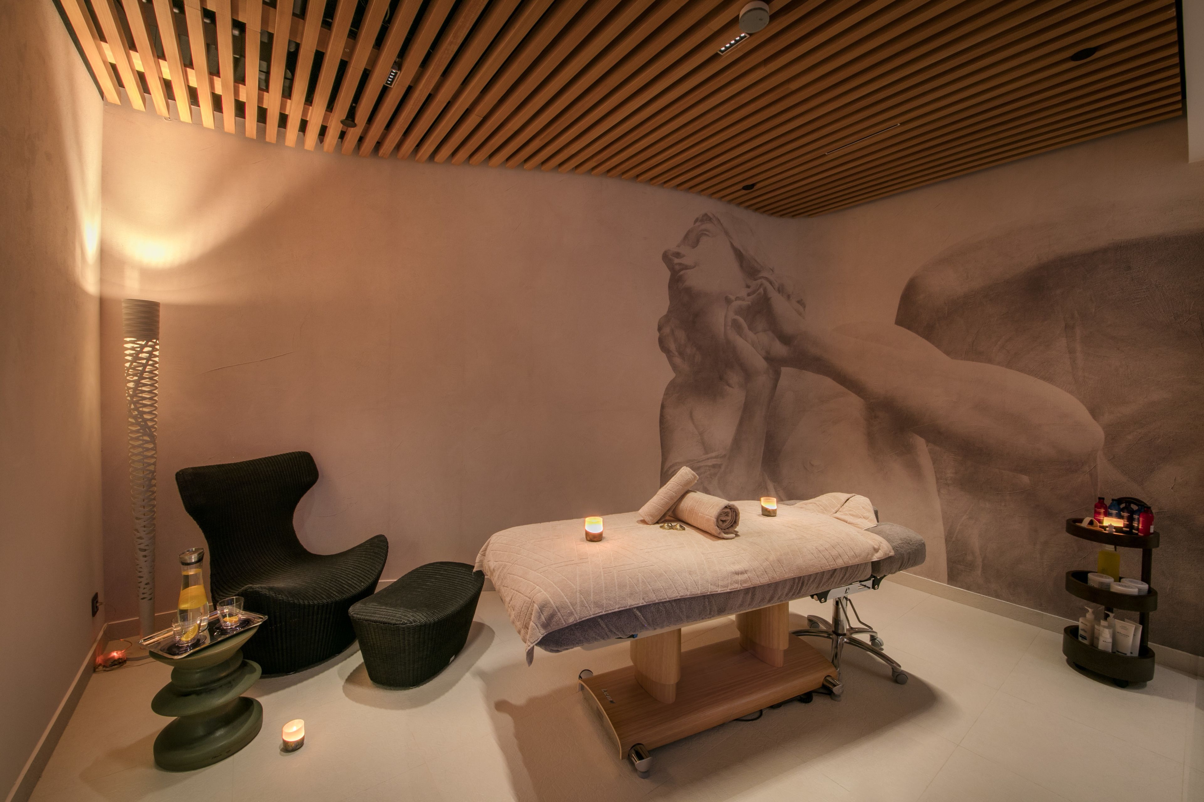 A spa room with a massage table, a black chair, a small table with candles, and a wall mural.