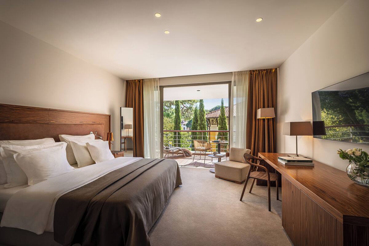 Ein Hotelzimmer mit einem großen Bett, einem Schreibtisch mit Lampe, einem Sessel und Blick ins Freie durch eine Glastür.