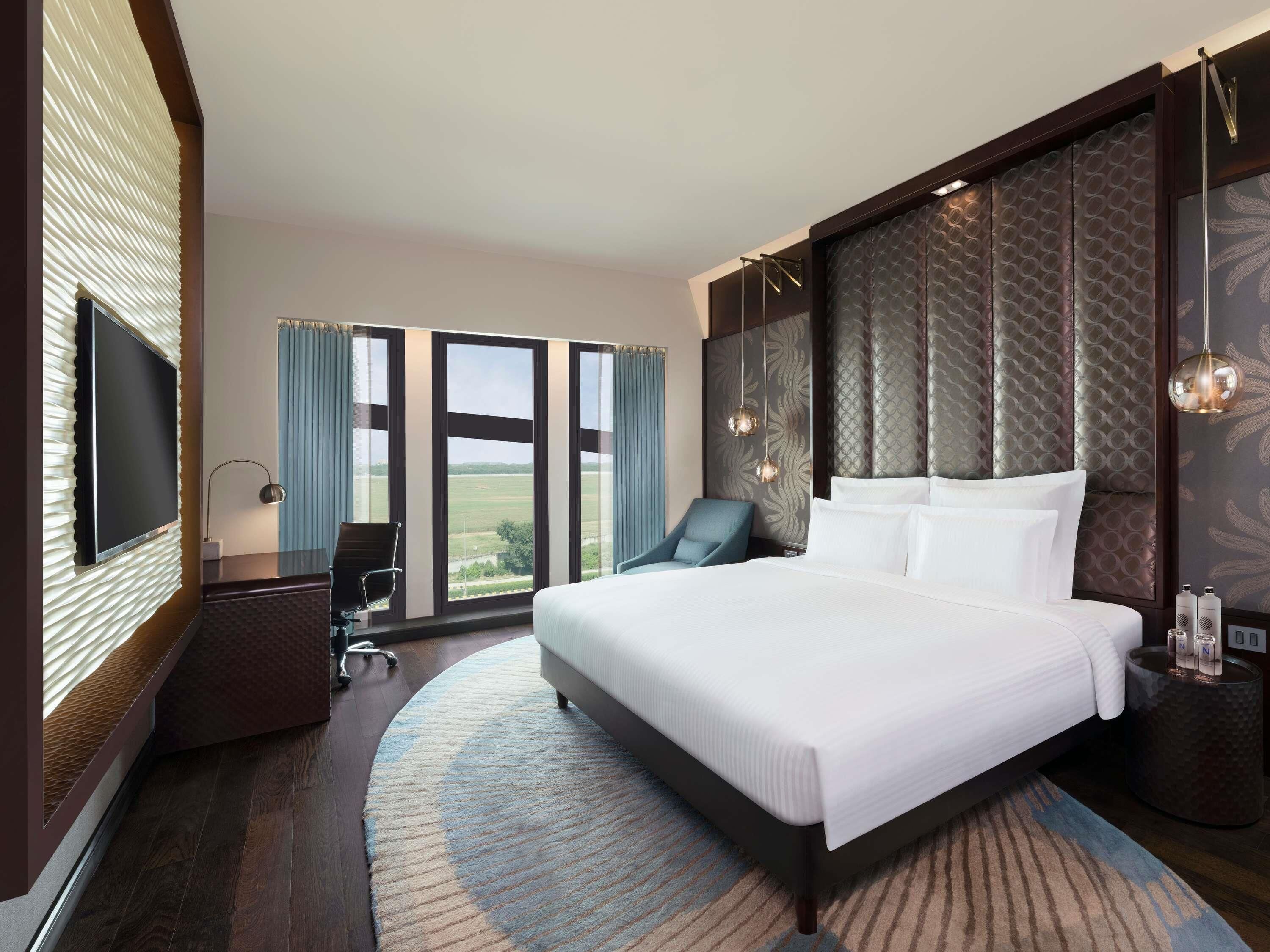 Une chambre d’hôtel moderne dotée d’un grand lit, d’un bureau avec une chaise, d’une télévision et offrant une vue sur l’extérieur grâce à de grandes fenêtres.