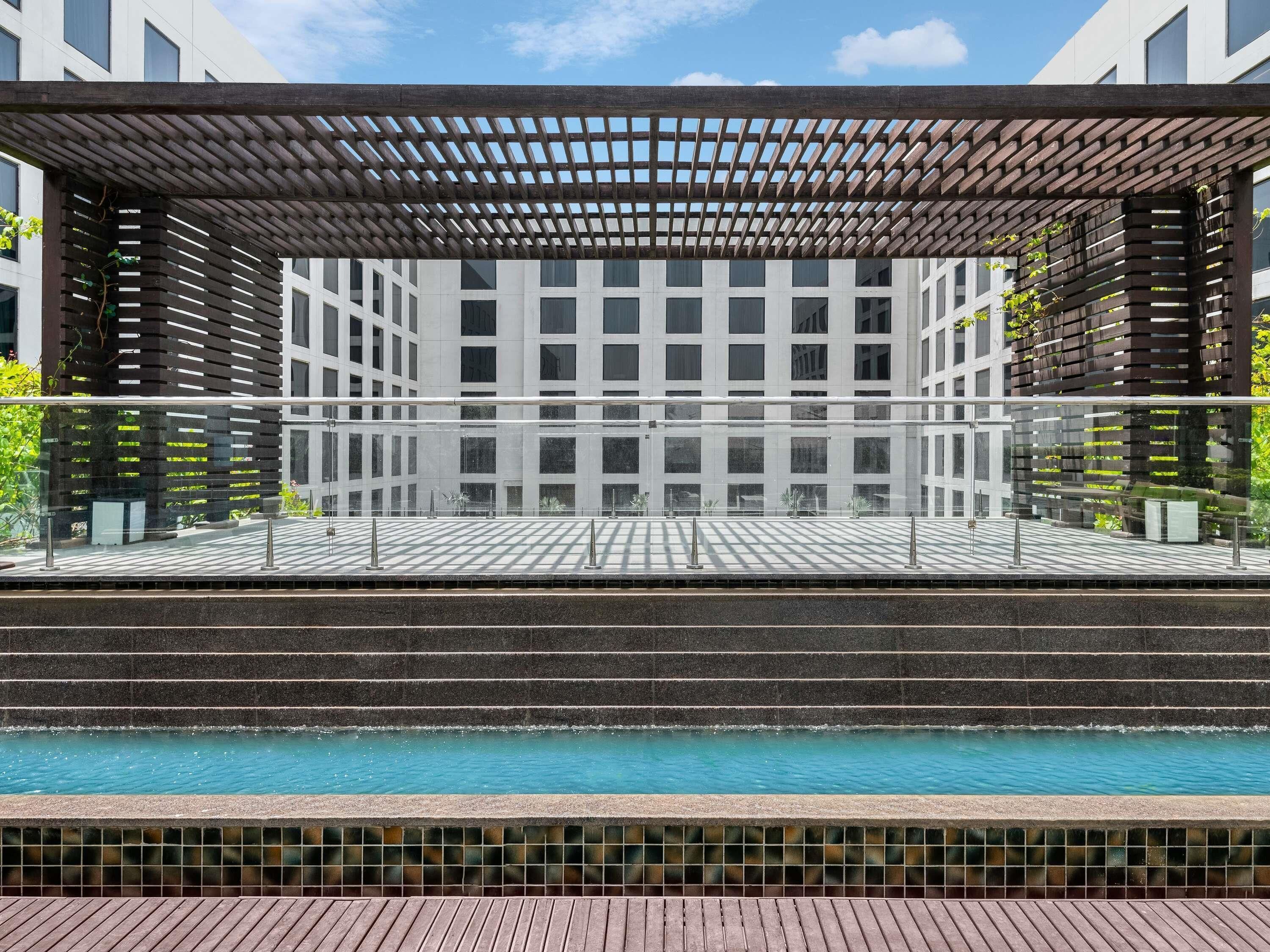 A pool area with a wooden pergola, a tiled deck, and a view of a modern building with a grid-like facade.