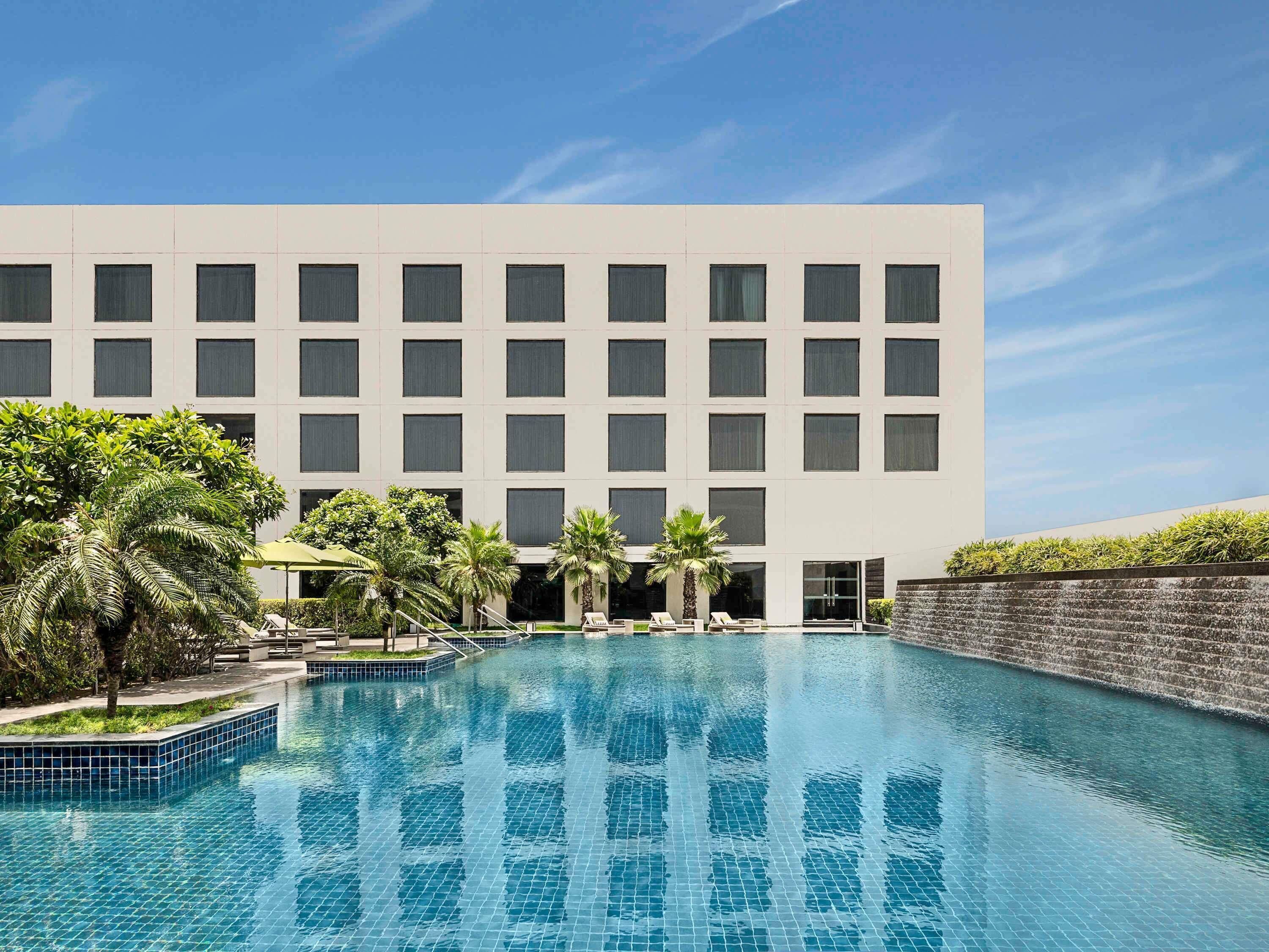 A modern hotel with a swimming pool, palm trees, and a clear blue sky.