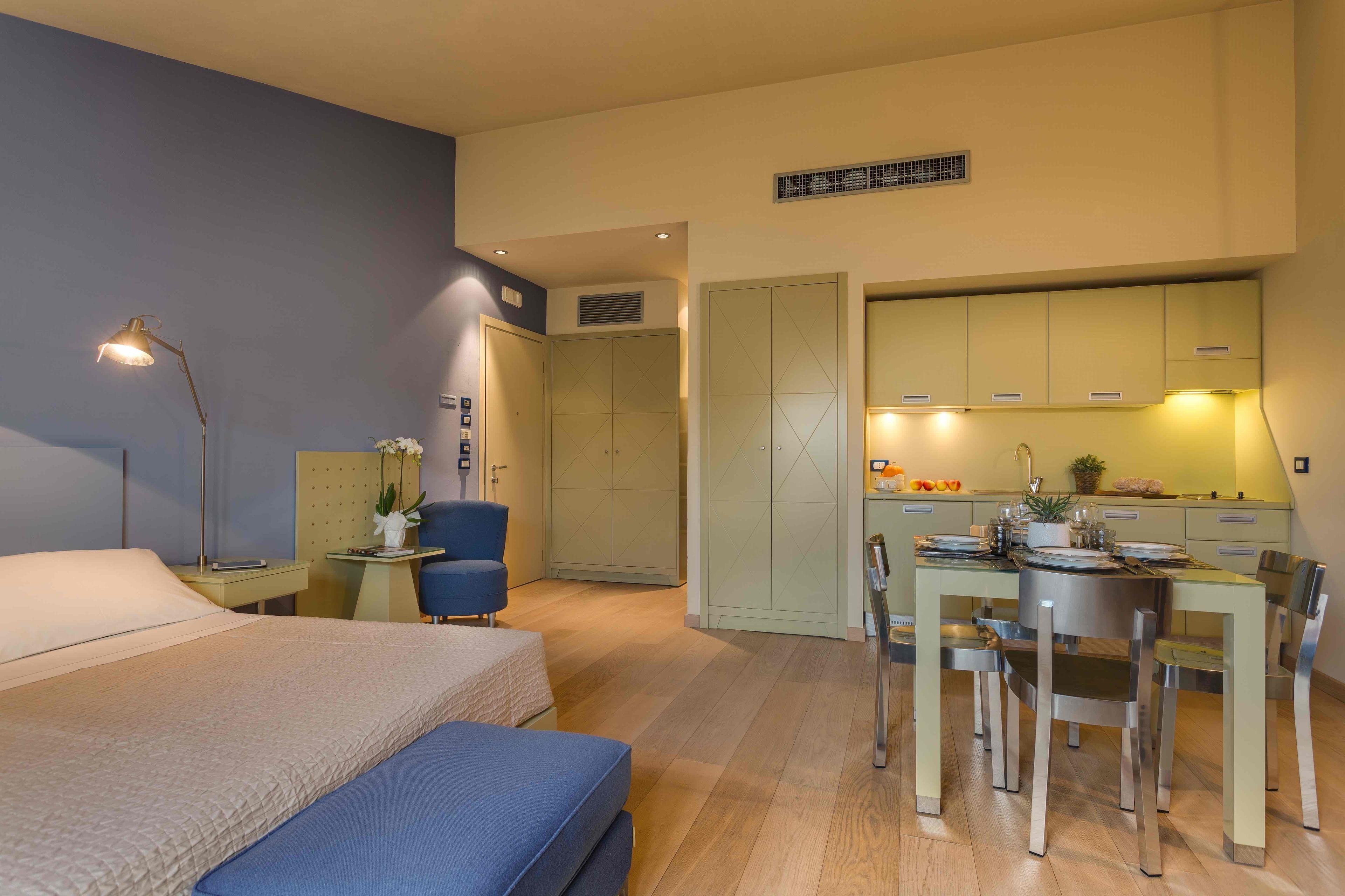 A hotel room with a bed, bedside table, lamp, chair, and a kitchenette area with a table, chairs, and a sink.