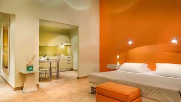 A bedroom with a bed, an orange bench, a small table with a lamp, and a kitchen area visible through an open doorway.