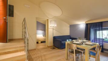 A modern hotel room with a wooden staircase, a blue sofa, a dining table set for four, and a large window with curtains.
