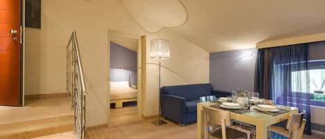 Una habitación de hotel moderna con una escalera de madera, un sofá azul, una mesa de comedor preparada para cuatro personas y un ventanal con cortinas.