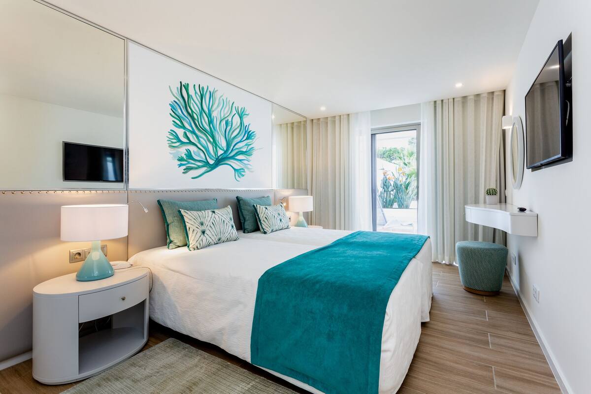 A modern hotel room with a large bed, a nightstand with a lamp, a wall-mounted TV, and a large window with curtains.