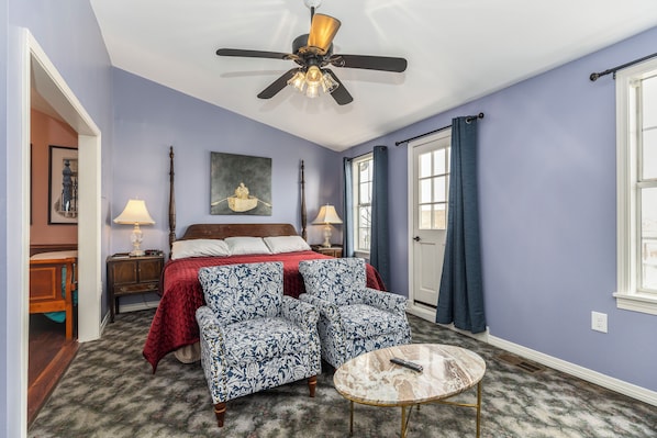 A bedroom with a bed, two chairs, a nightstand, a lamp, a ceiling fan, and a window with curtains.