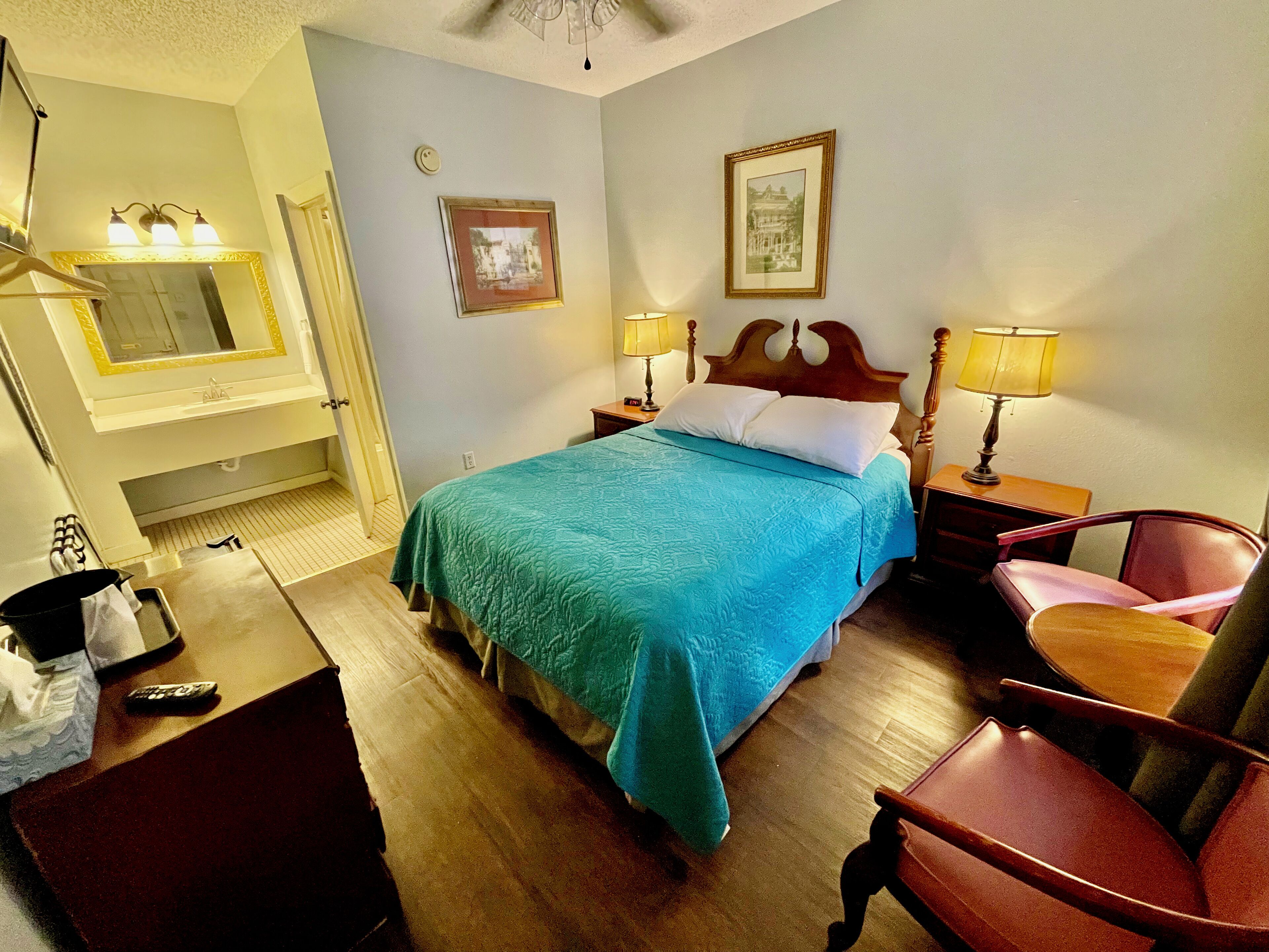 A hotel room with a bed, two bedside lamps, a chair, a desk, a mirror, and two framed pictures on the wall.