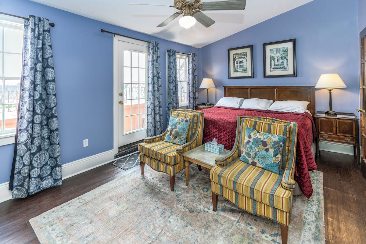 Um quarto com cama, duas cadeiras, mesa de cabeceira e ventilador de teto.