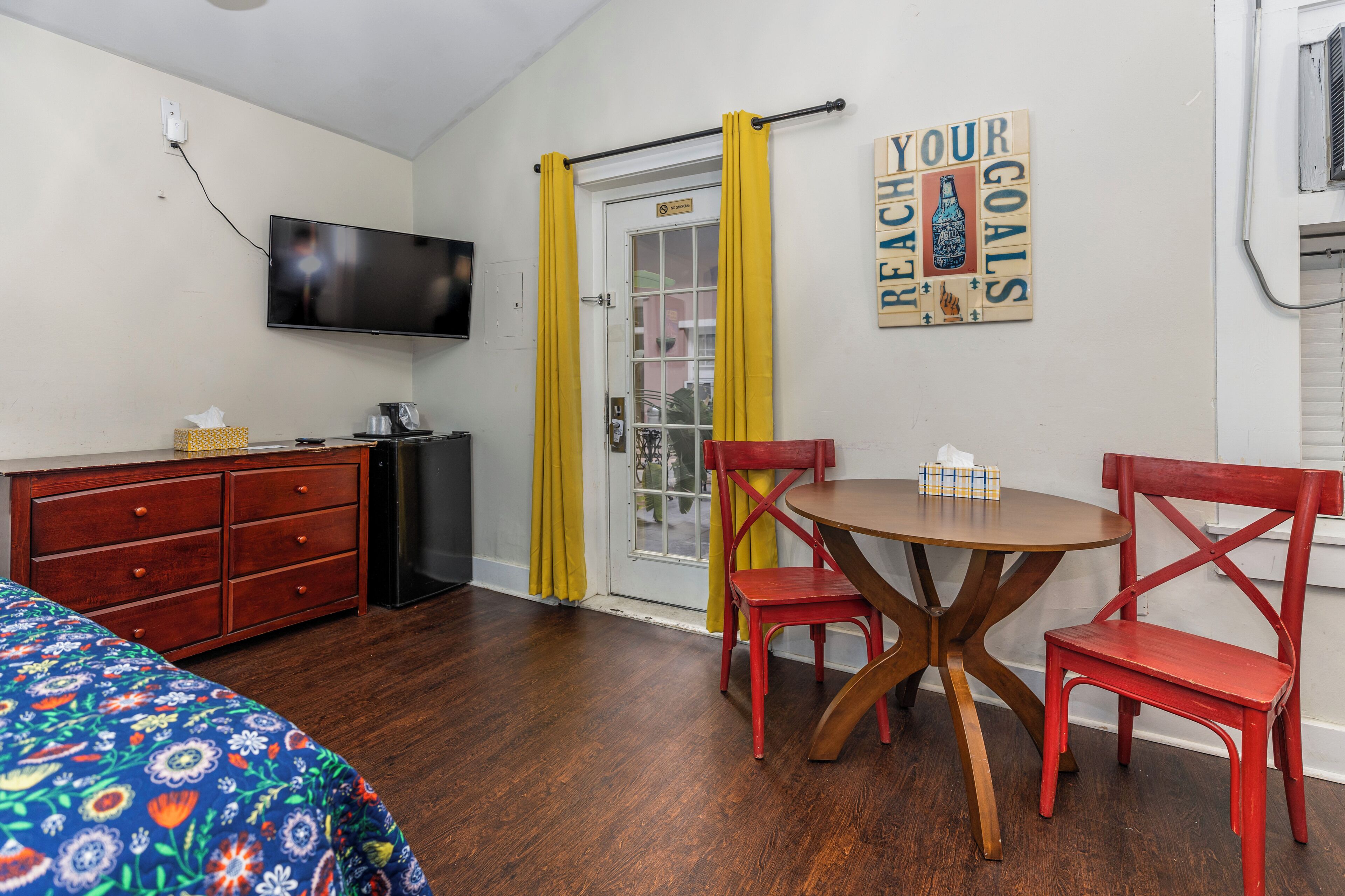 Een kamer met een bed, een televisie, een bureau en een stoel. Er is een deur met gele gordijnen.