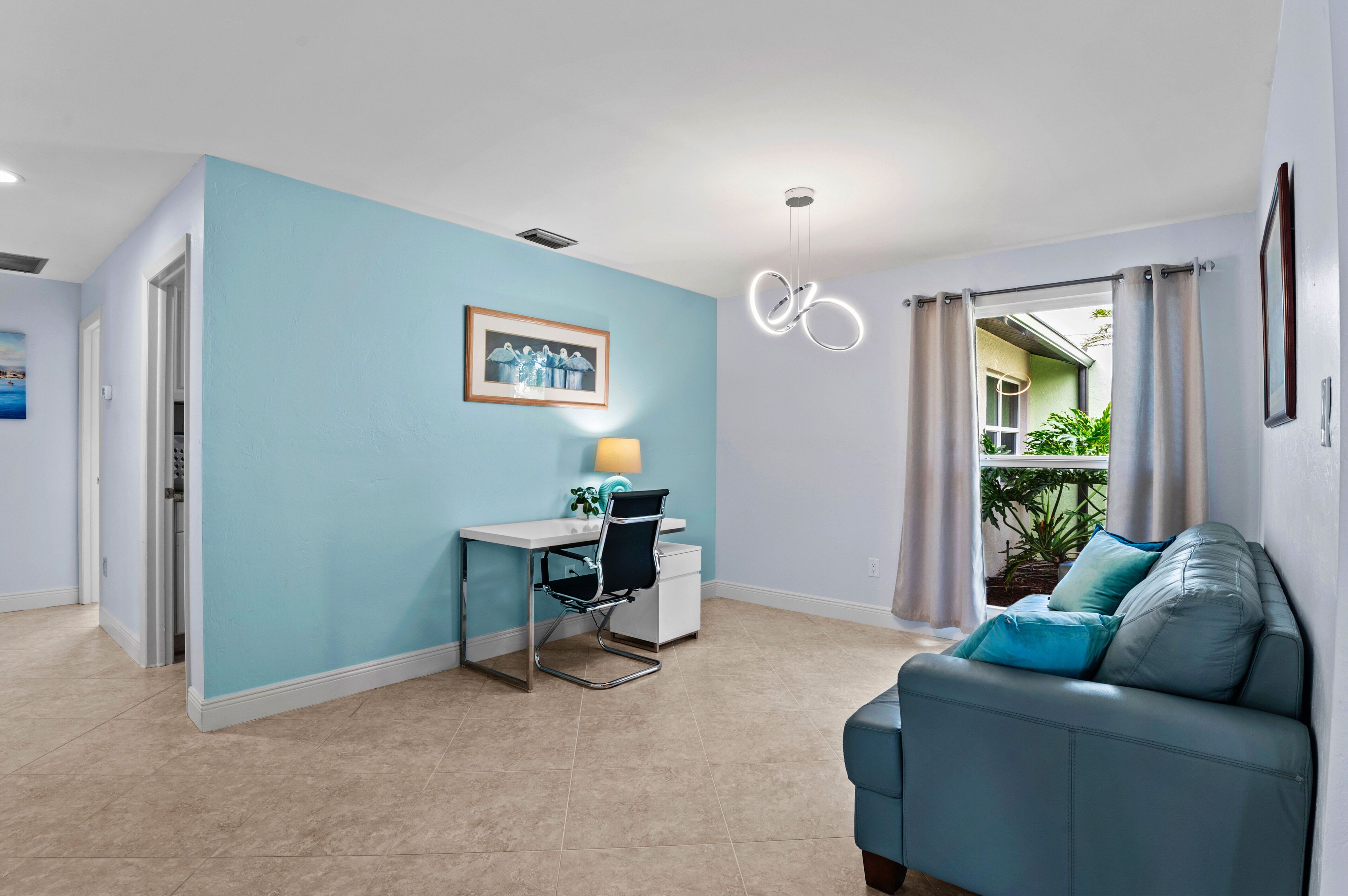 Un salon moderne avec un mur d’accent bleu, un bureau avec une chaise, un canapé avec des coussins et une vue sur l’extérieur.