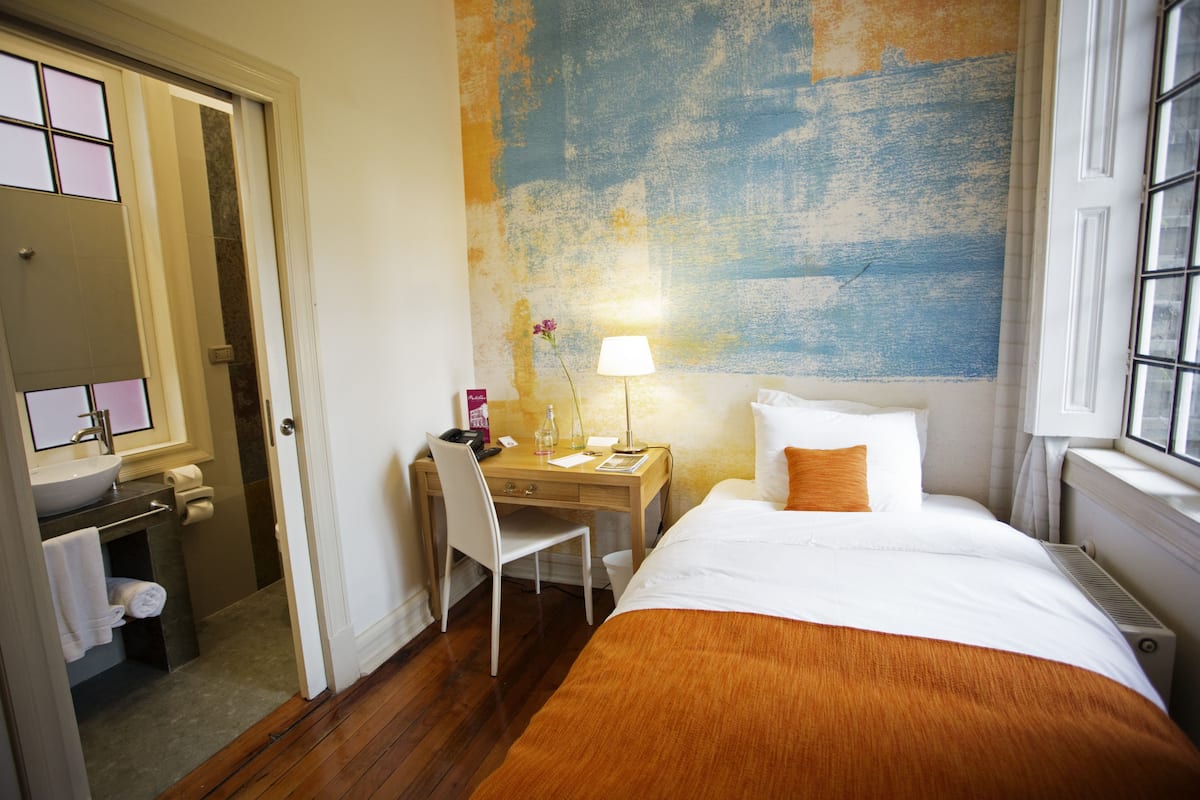 A hotel room with a bed, a desk, a chair, a lamp, and a bathroom with a sink and mirror.