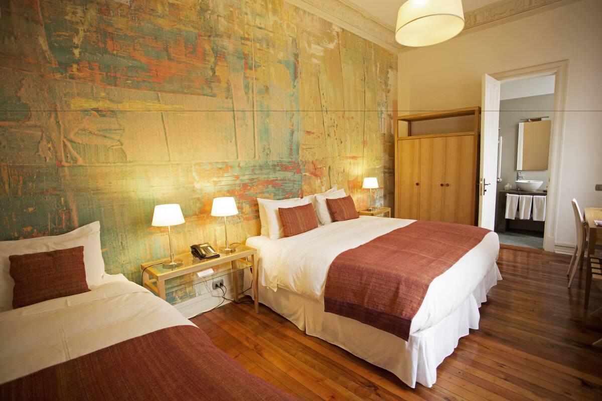 A hotel room with two beds, wooden flooring, a large painting on the wall, and a bathroom visible through an open door.