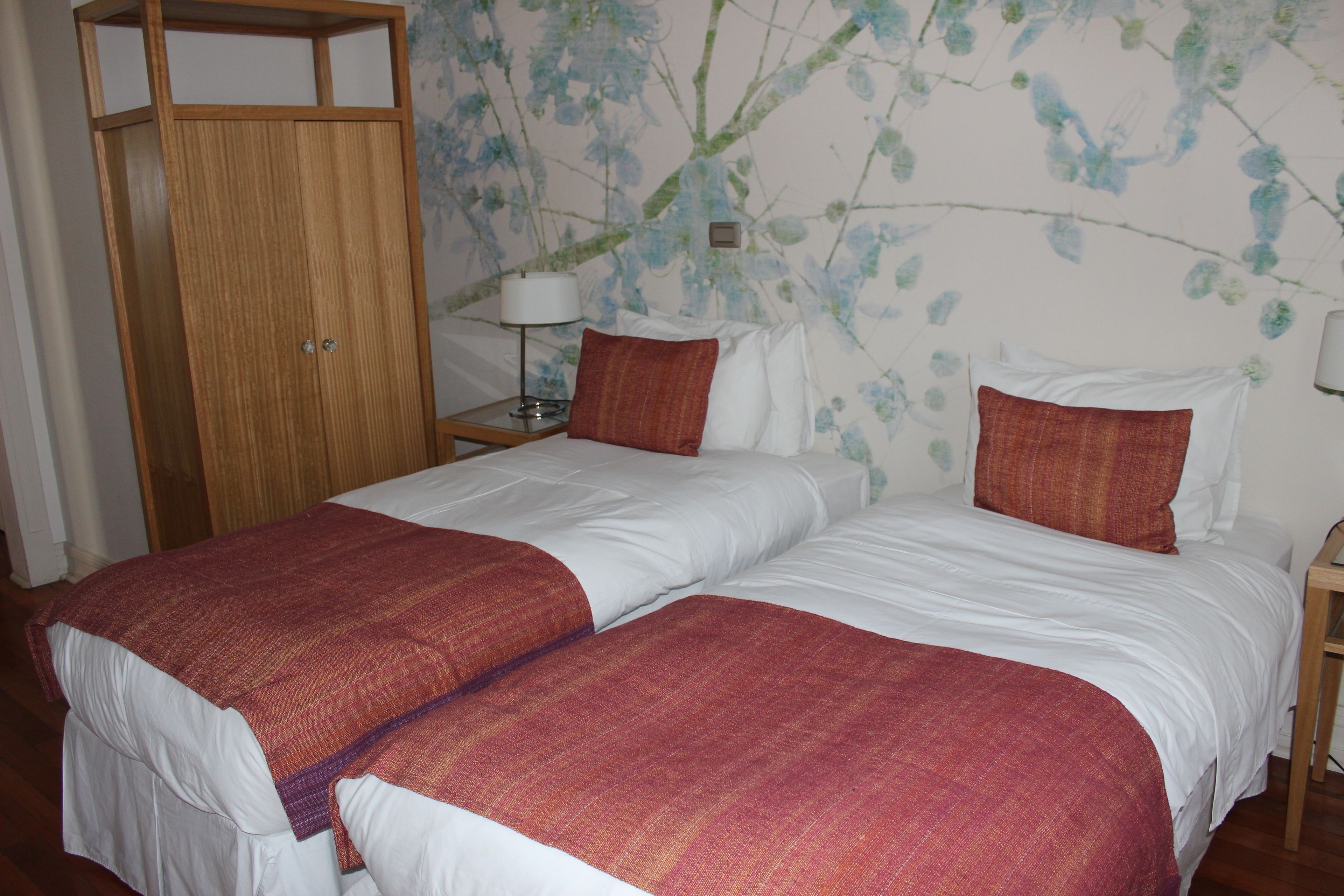 A double bed with white and red bedding, a wooden wardrobe, and a wall with floral wallpaper.