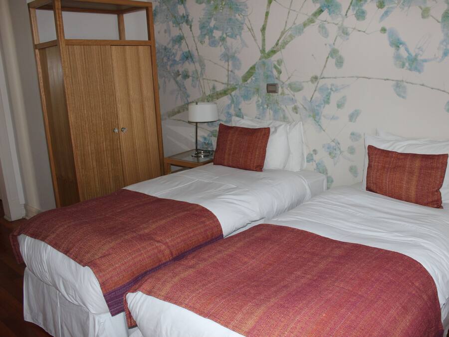 A double bed with white and red bedding, a wooden wardrobe, and a wall with floral wallpaper.