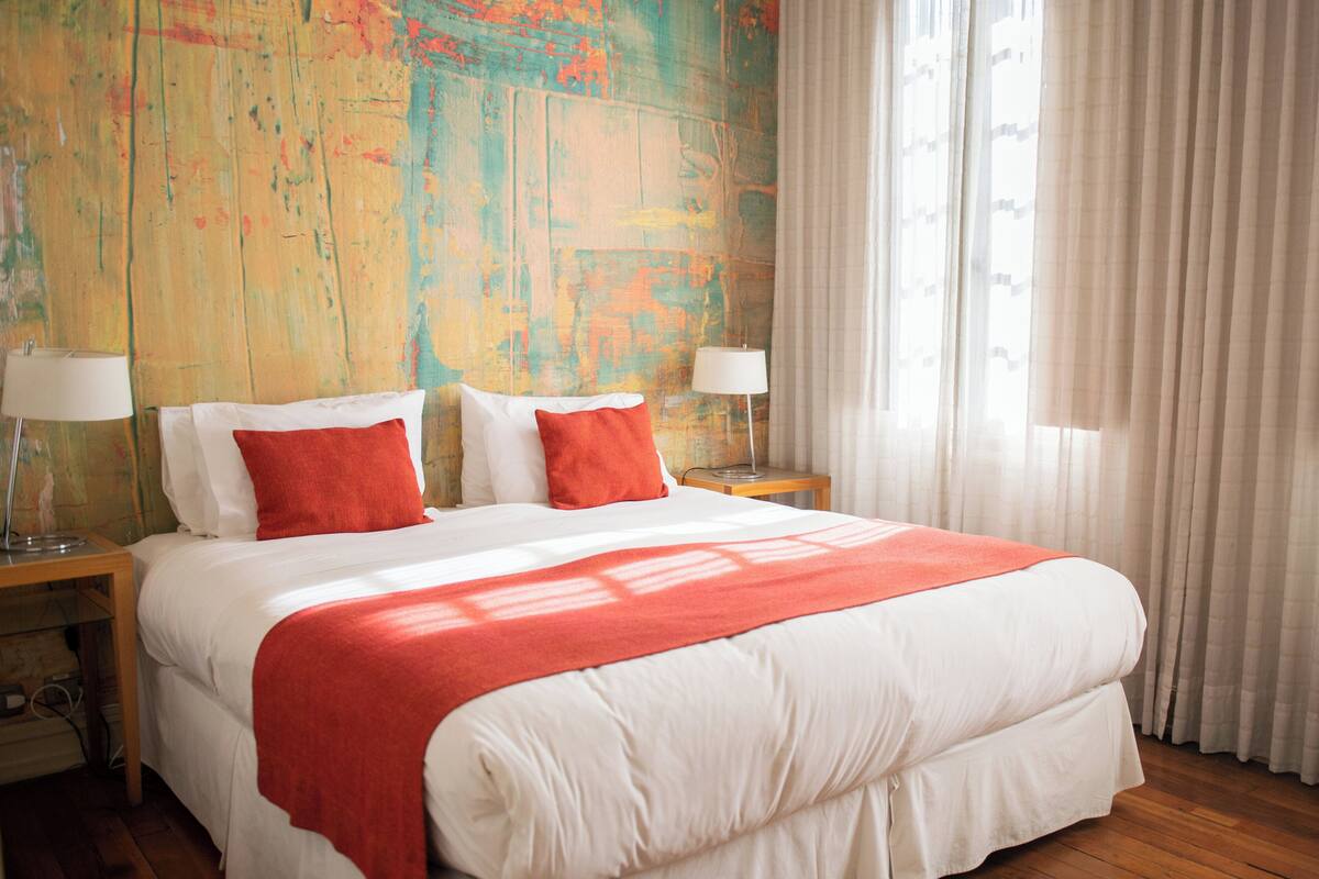 A large bed with white and red bedding, two bedside tables with lamps, and a colorful abstract wall.
