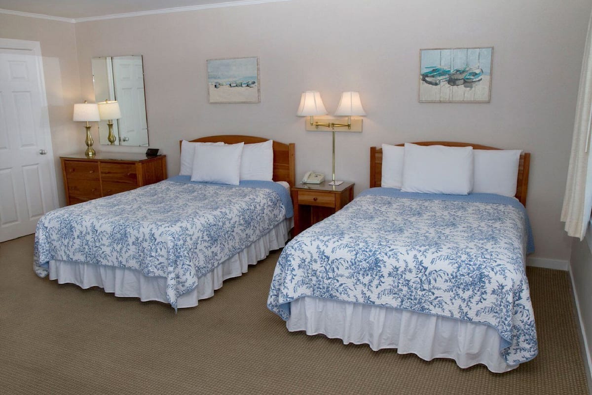 A hotel room with two single beds, a wooden dresser, a nightstand with a lamp, and two wall-mounted lamps.