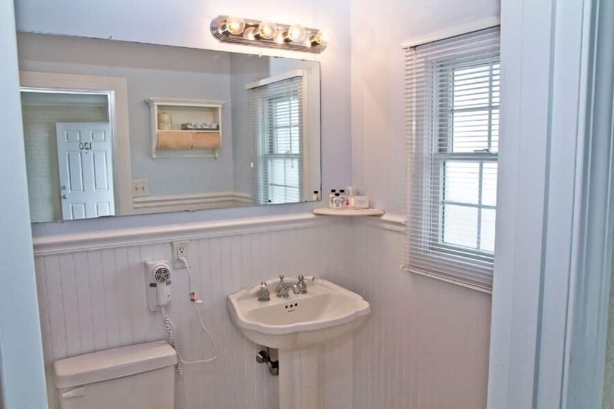 A bathroom with a pedestal sink, a mirror with lights, a toilet, and a window with blinds.