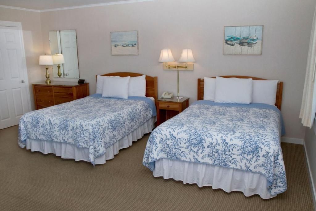 A hotel room with two beds, a wooden dresser, a mirror, a lamp, and artwork on the walls.