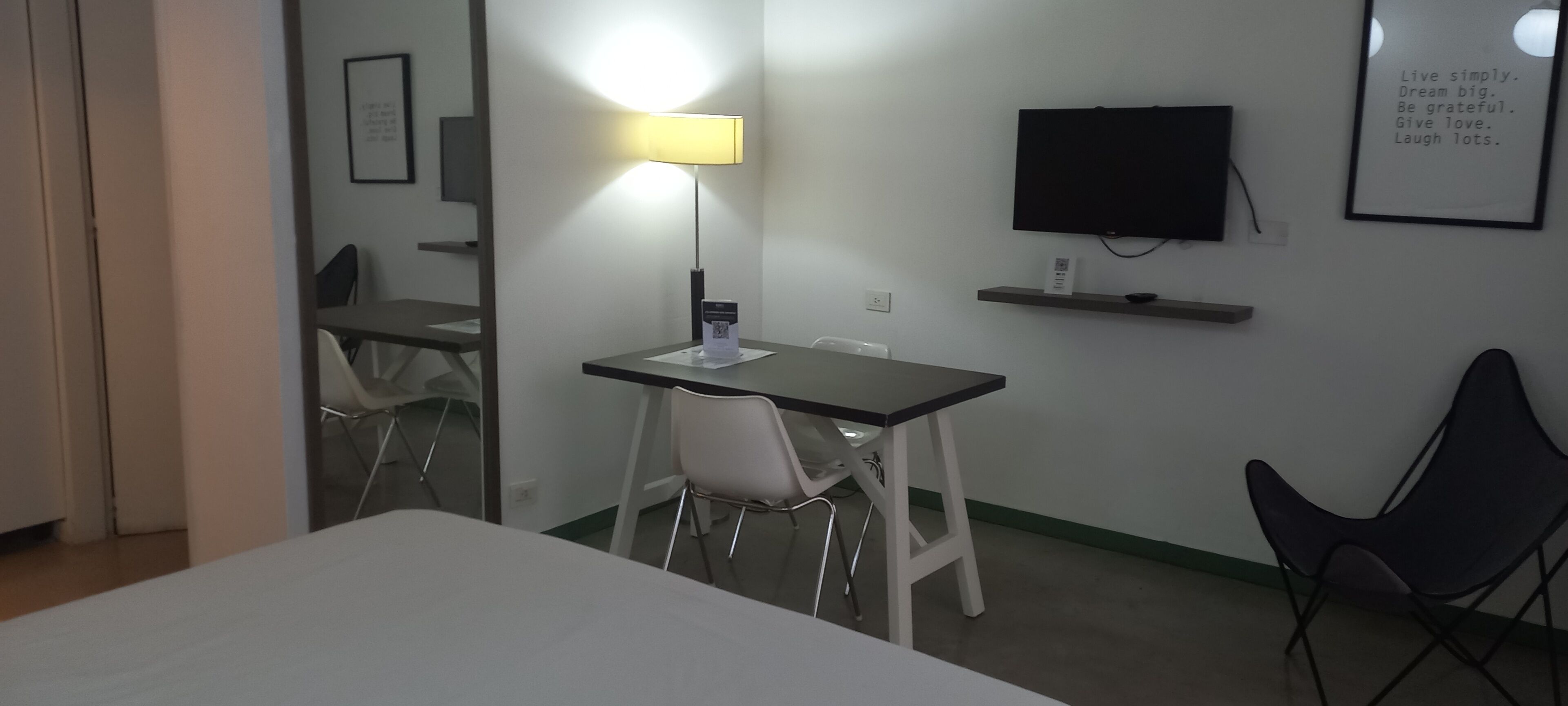 Une chambre d’hôtel comprenant un lit, un bureau avec une chaise, une lampe sur pied, un téléviseur et une affiche motivante accrochée au mur.