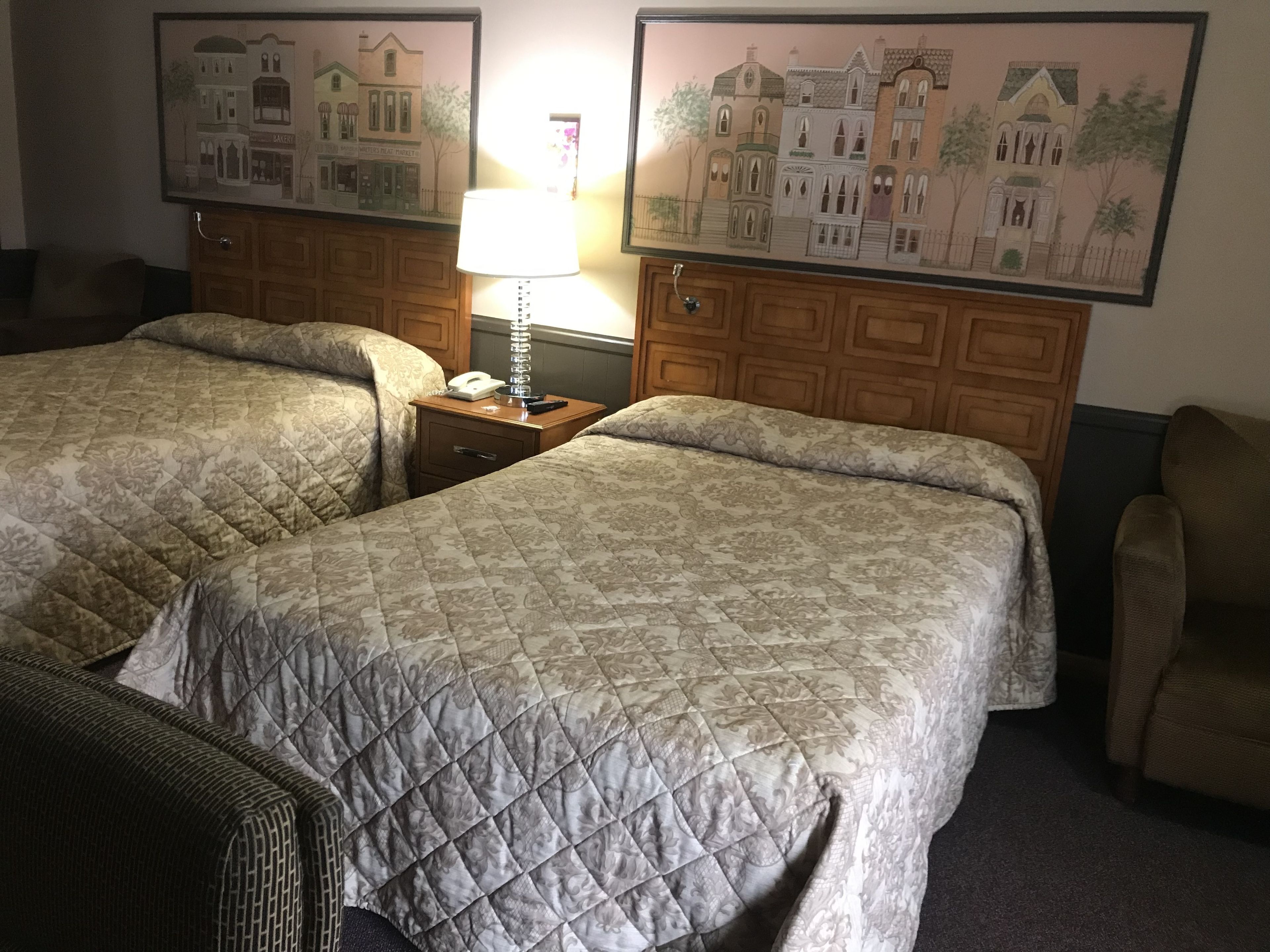 A hotel room with two beds, a nightstand with a lamp, a chair, and two framed pictures on the wall.