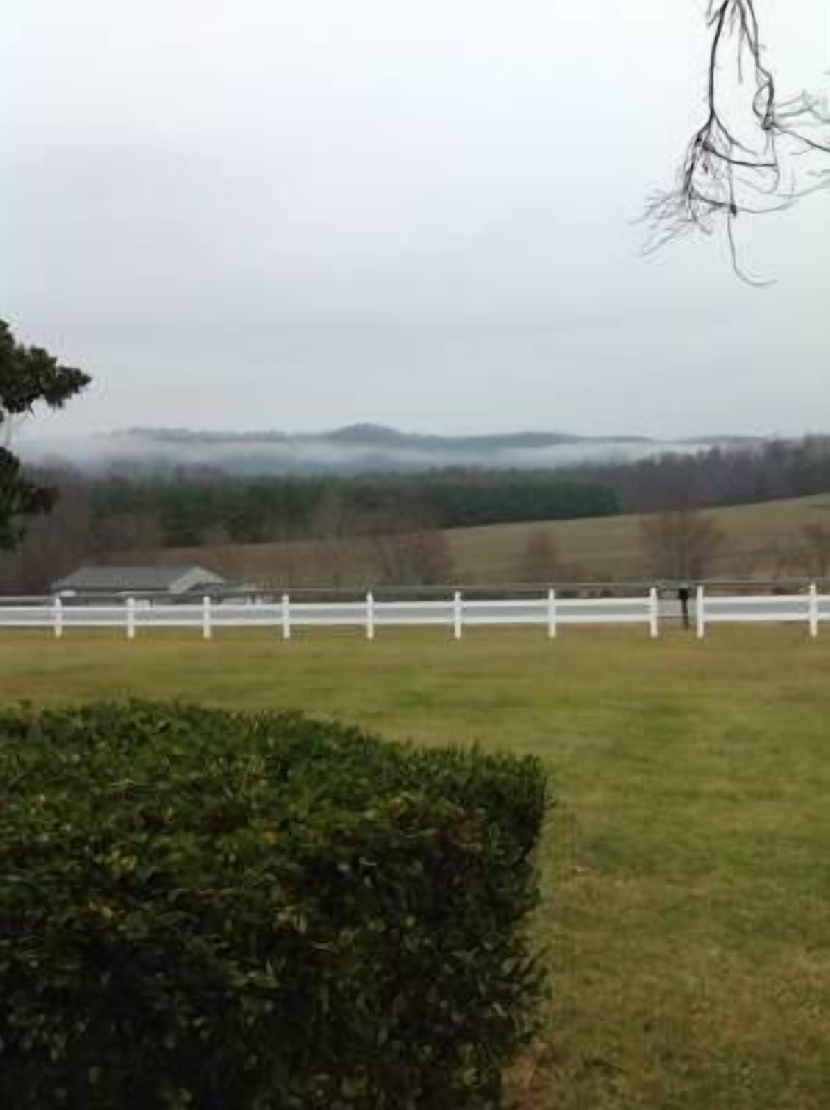 A stable with a white fence, a hedge, and a distant view of a landscape with trees and hills.