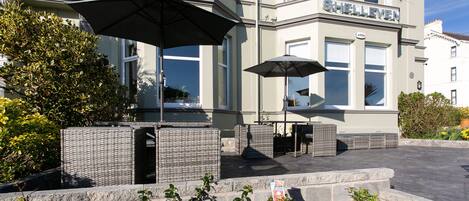 A building with the name "SHELLVEN" on the facade, outdoor seating with umbrellas, and a garden area with plants.