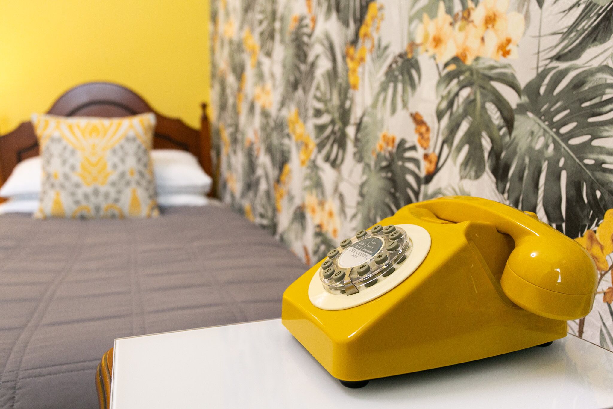 A yellow rotary phone on a white table in a hotel room with a bed, a patterned wall, and a wooden headboard.
