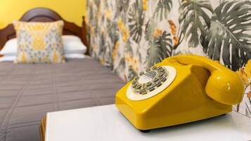 A yellow rotary phone on a white table in a hotel room with a bed, a patterned wall, and a wooden headboard.