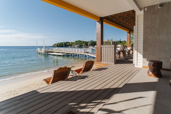 A wooden deck with two chairs and a table overlooking a beach and pier. - The Beach House Boutique Hotel (Roatan)