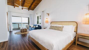 A bedroom with a large bed, wooden ceiling beams, a ceiling fan, a view of the ocean through a sliding door, and a small round table with chairs.