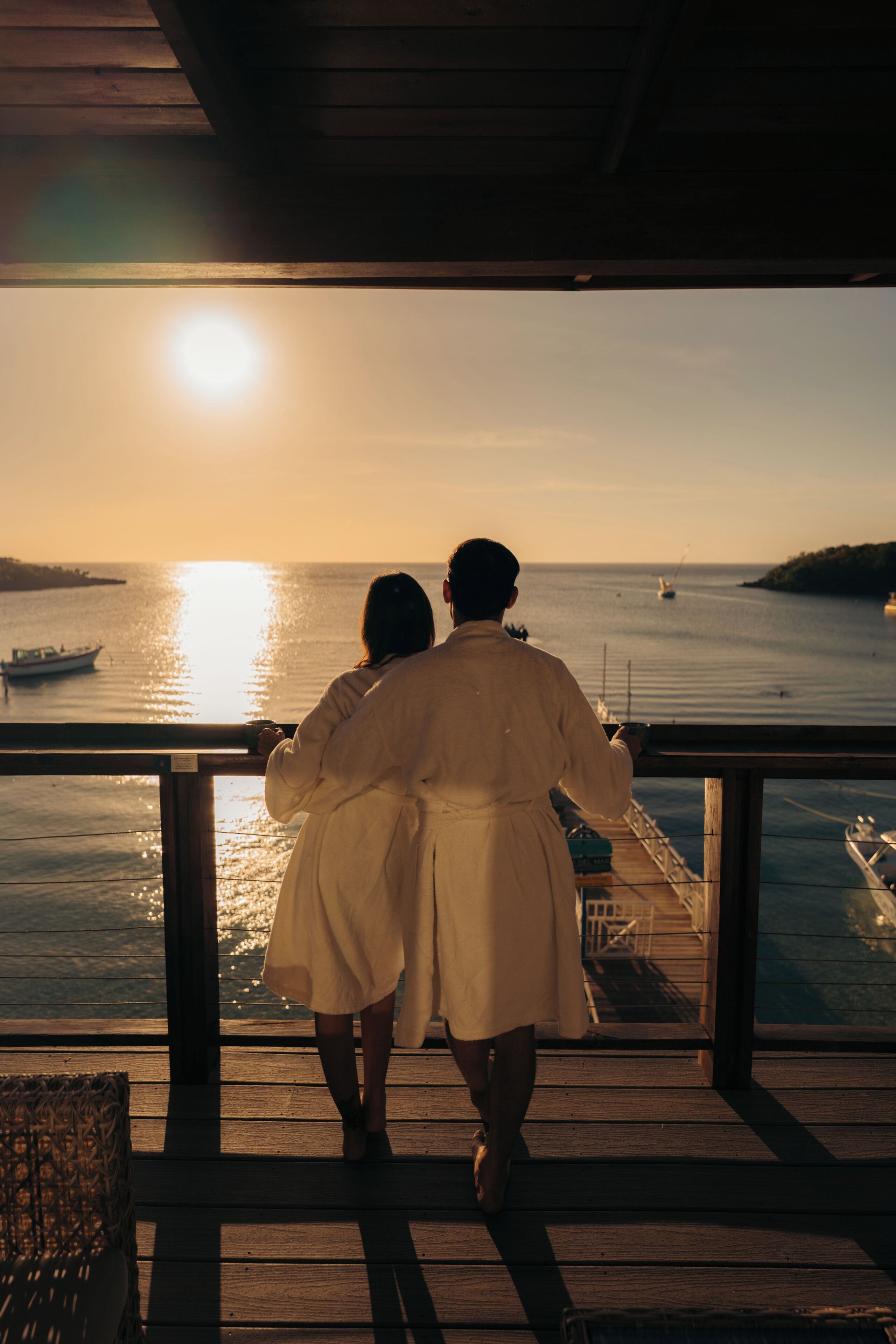 Una pareja está de pie en una terraza de madera con vista a un cuerpo de agua y barcos, durante el atardecer.