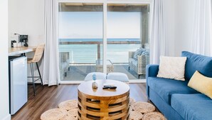 Une pièce moderne avec un plafond en bois, une table ronde en bois surmontée d’un tapis tissé, un canapé bleu et une vue sur la mer, visible à travers de grandes portes vitrées.