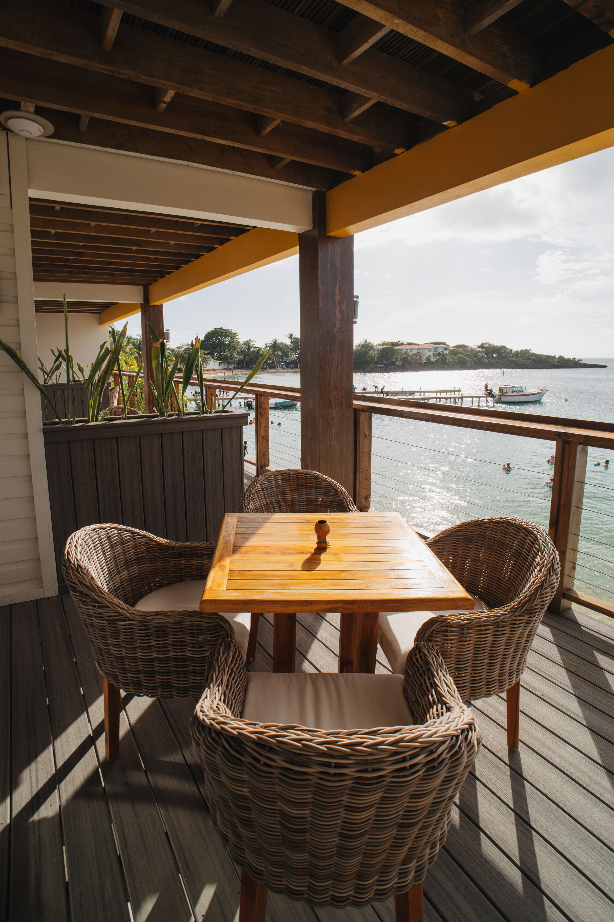 Une terrasse en bois avec une table et des chaises en osier, donnant sur une étendue d’eau où l’on peut voir des bateaux et des gens.