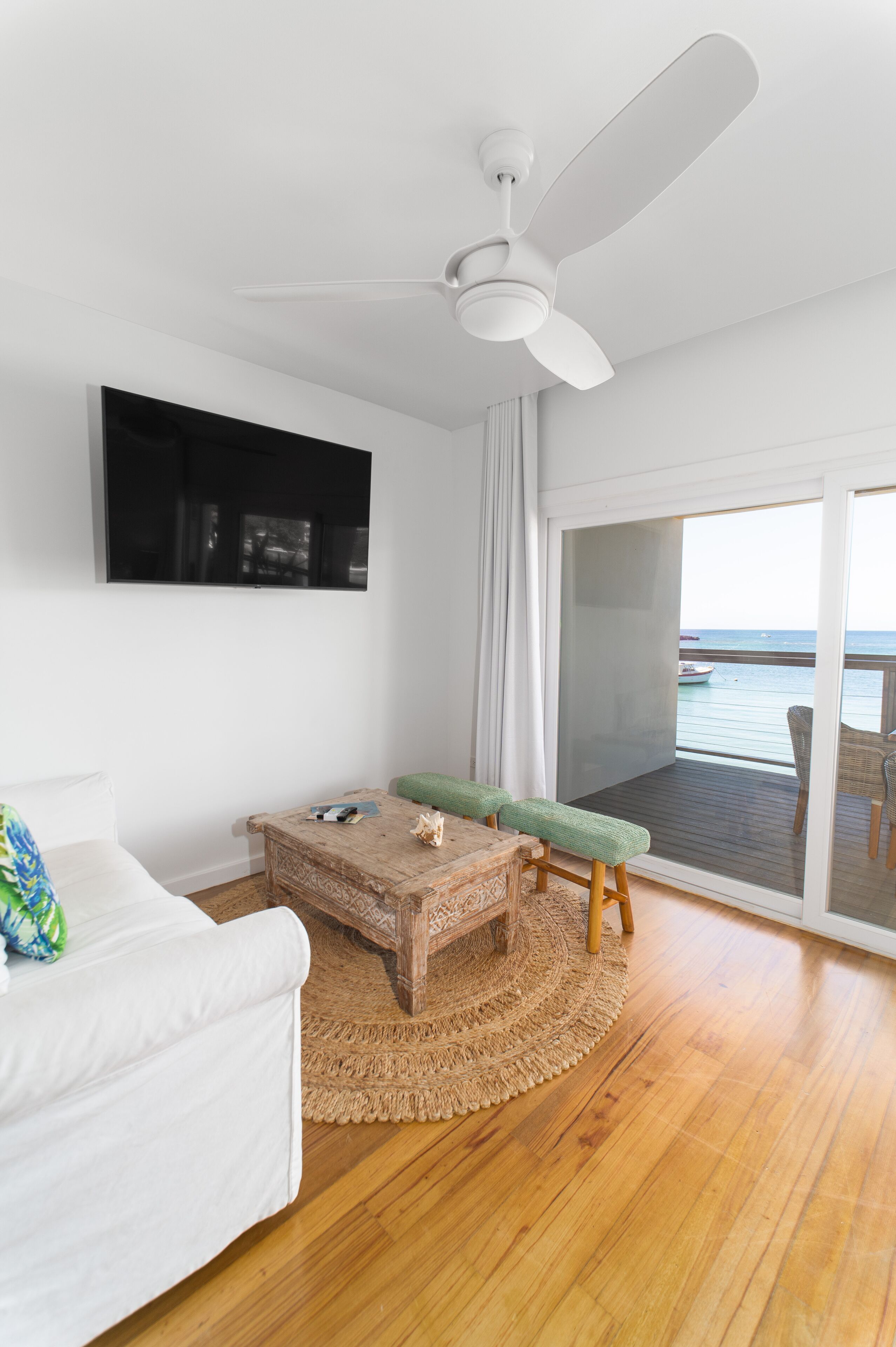 A room with a ceiling fan, a flat-screen TV, a wooden coffee table, a green bench, a white sofa, and a view of the sea through a sliding glass door.