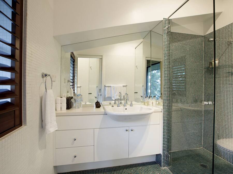 Un baño moderno con un espejo grande, muebles blancos, cabina de ducha de vidrio y vistas al exterior.