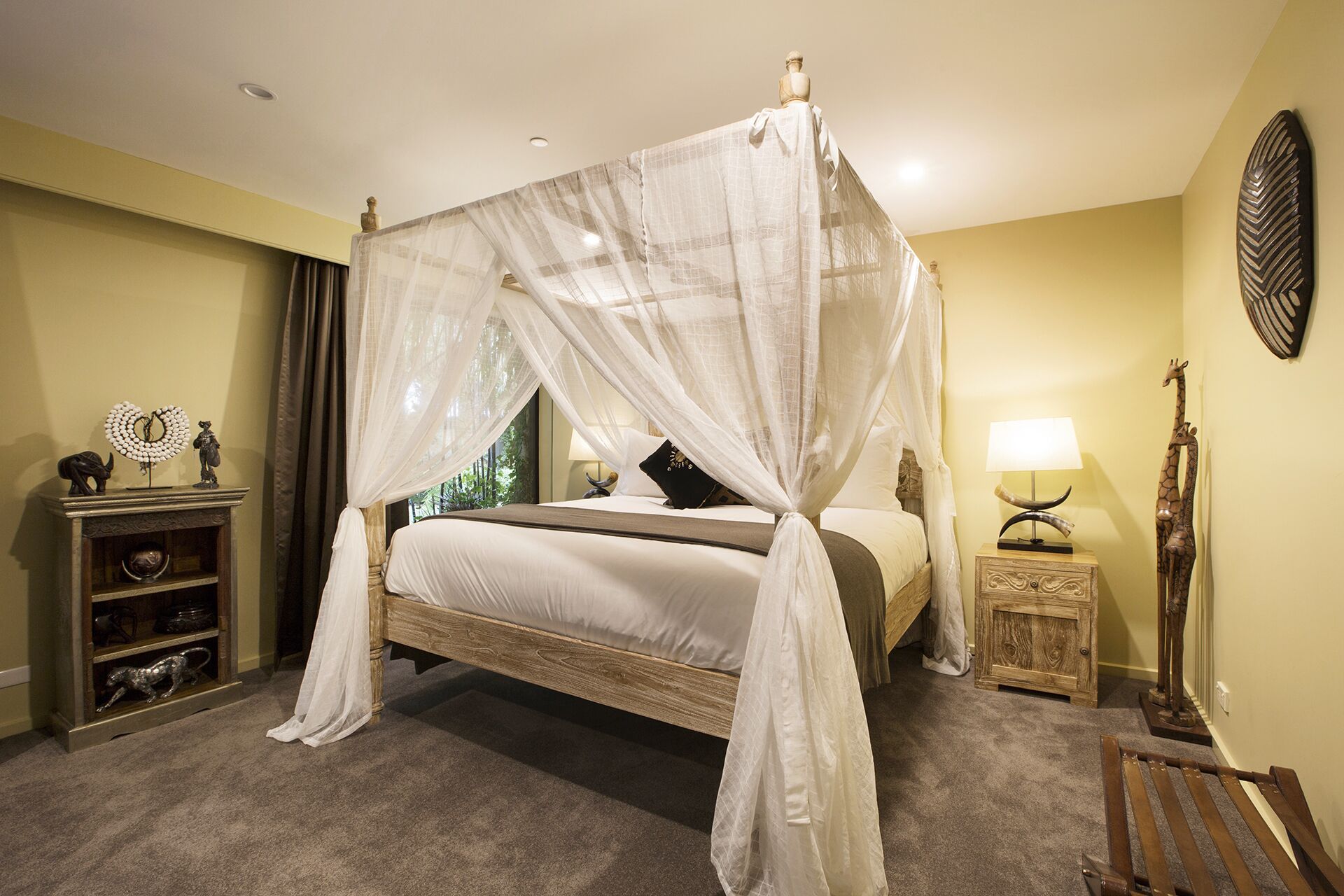 A four-poster canopy bed with a wooden frame and white bedding, a bedside table with a lamp, and a giraffe sculpture on the wall.