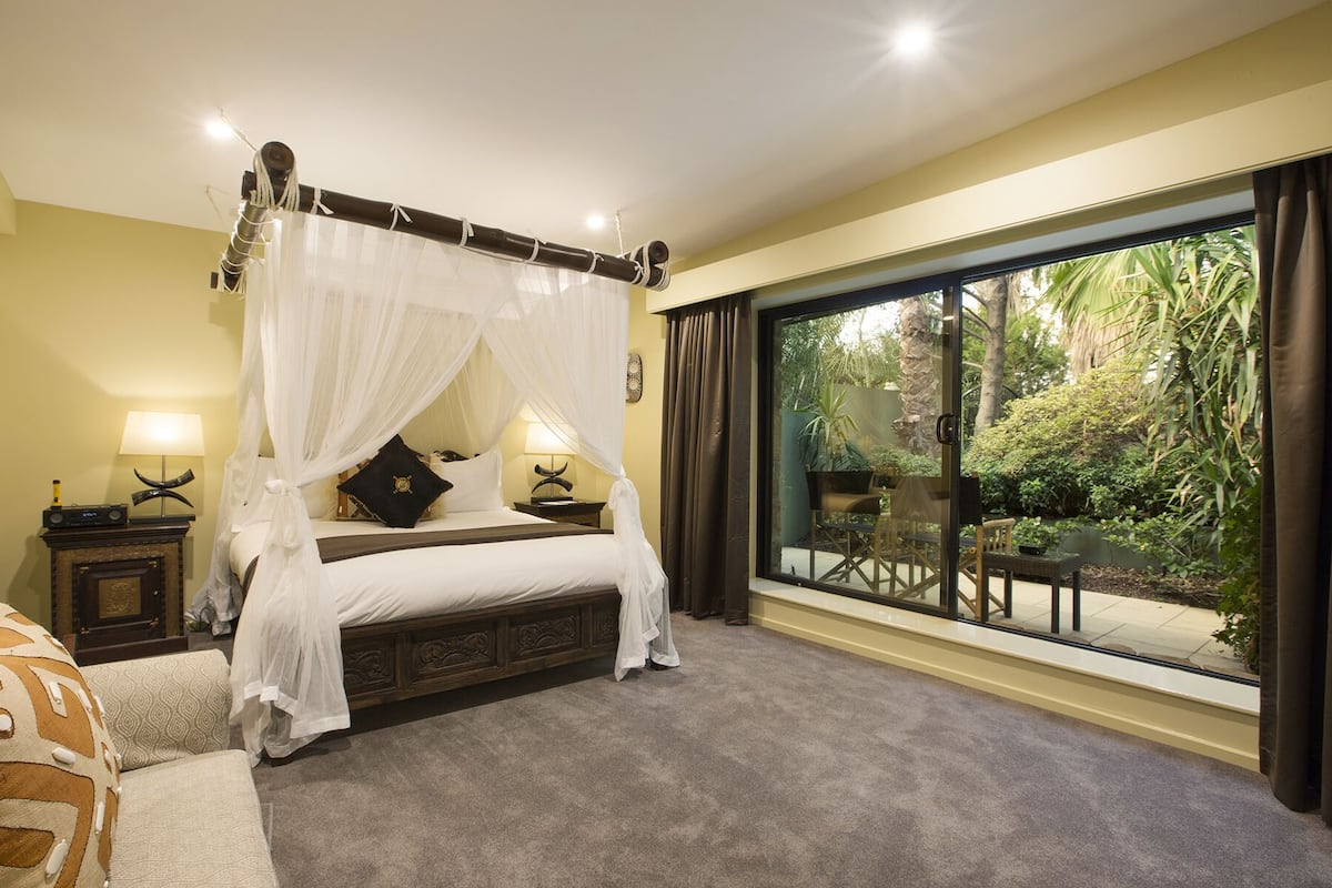 Ein Schlafzimmer mit einem Himmelsbett, zwei Nachttischlampen, einem großen Fenster mit Blick auf Grünflächen und einem kleinen Tisch mit Stühlen davor.