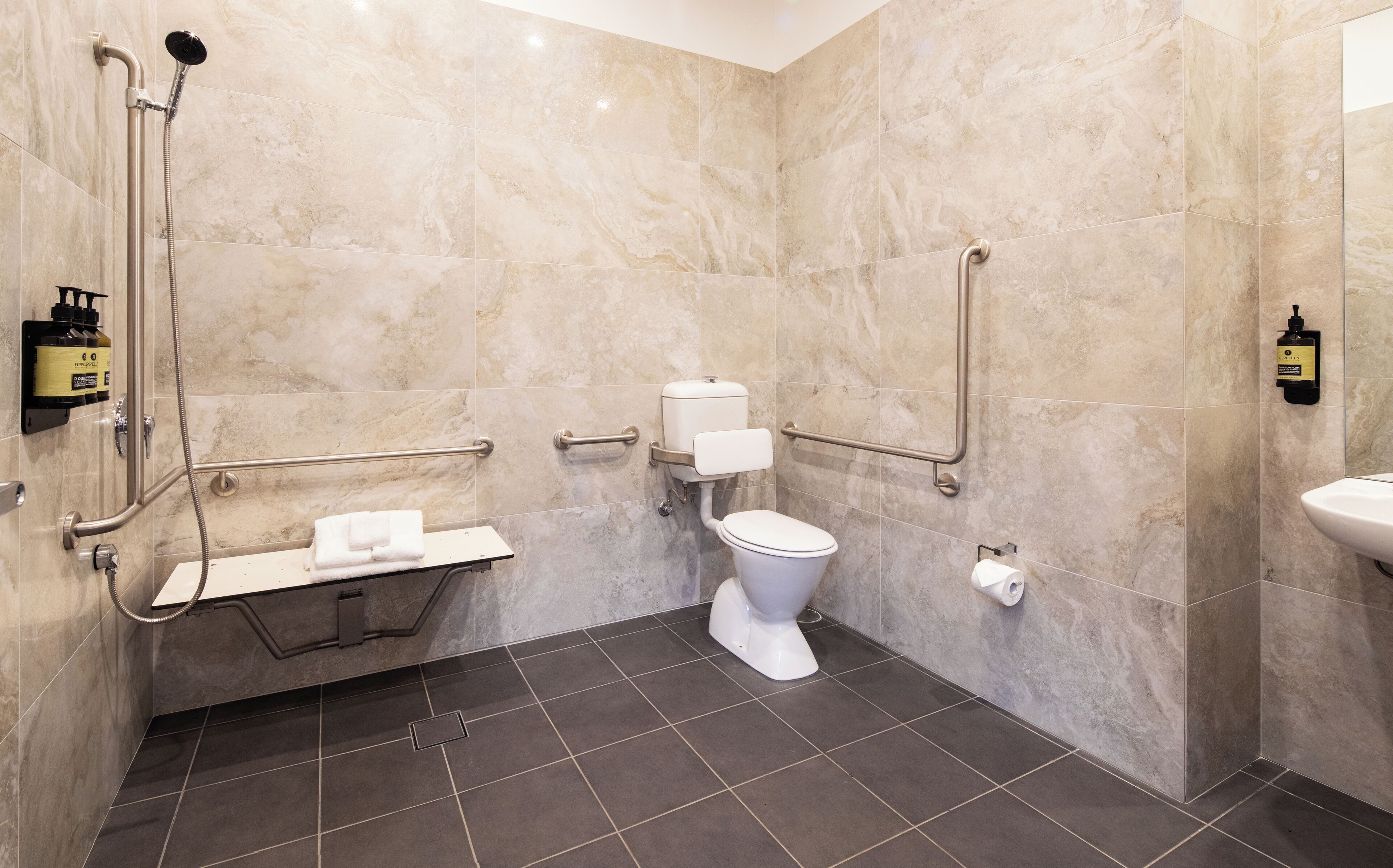 A modern bathroom with a shower, toilet, and sink, equipped with grab bars and a hand dryer.