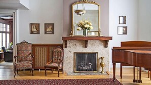 A spacious living room with a grand piano, a fireplace with a mirror above, and three upholstered chairs. - Shadow Lawn Inn (Rothesay)