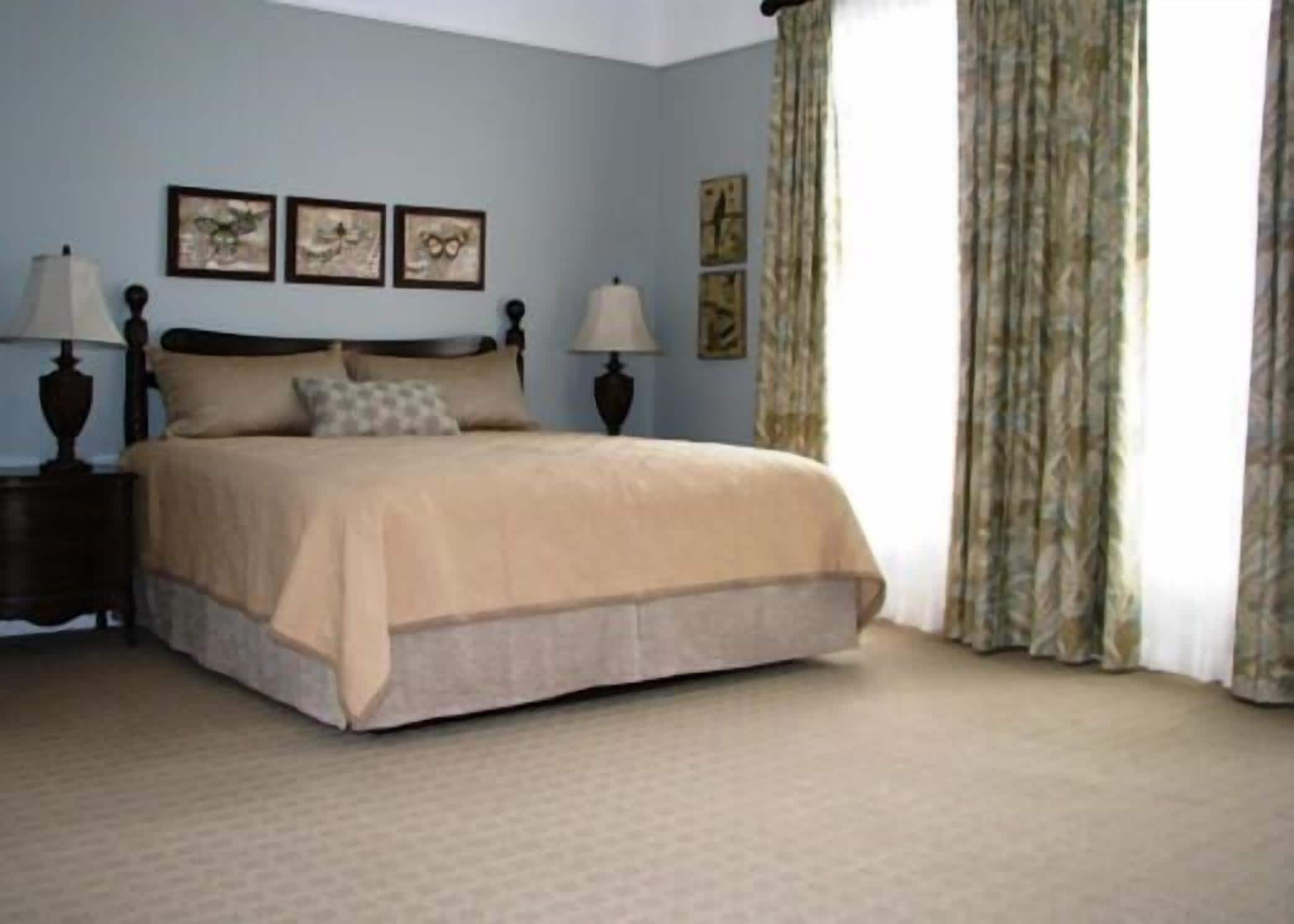 A neatly made bed with a beige bedspread in a room with light blue walls, two framed pictures on the wall, and a window with curtains.