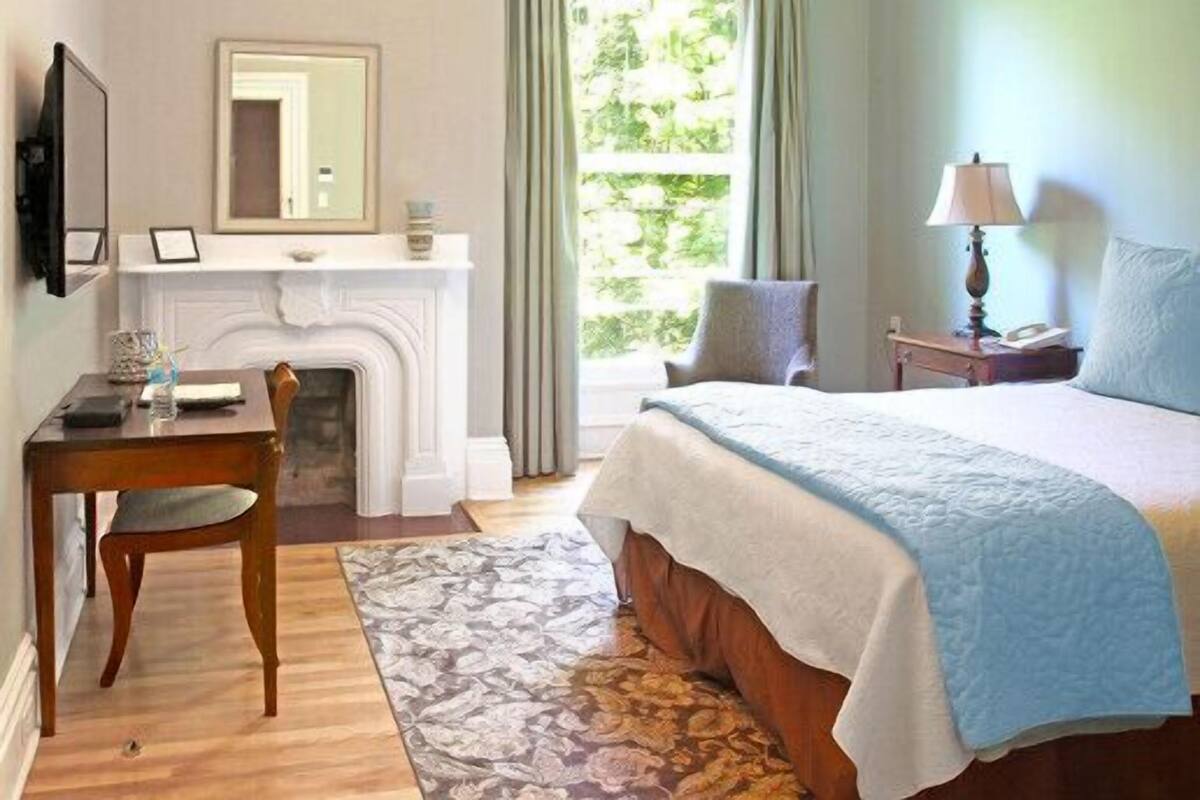 Un dormitorio con cama, escritorio, silla, chimenea, ventana con cortinas y mesita de noche con lámpara.