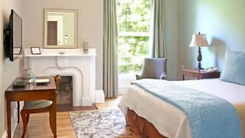 A bedroom with a bed, a desk, a chair, a fireplace, a window with curtains, and a nightstand with a lamp.