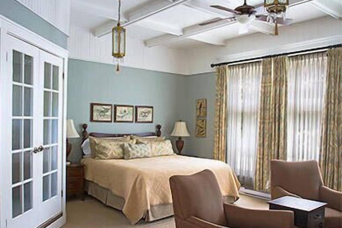 Una habitación con cama, silla, mesa, ventilador de techo y ventana con cortinas.