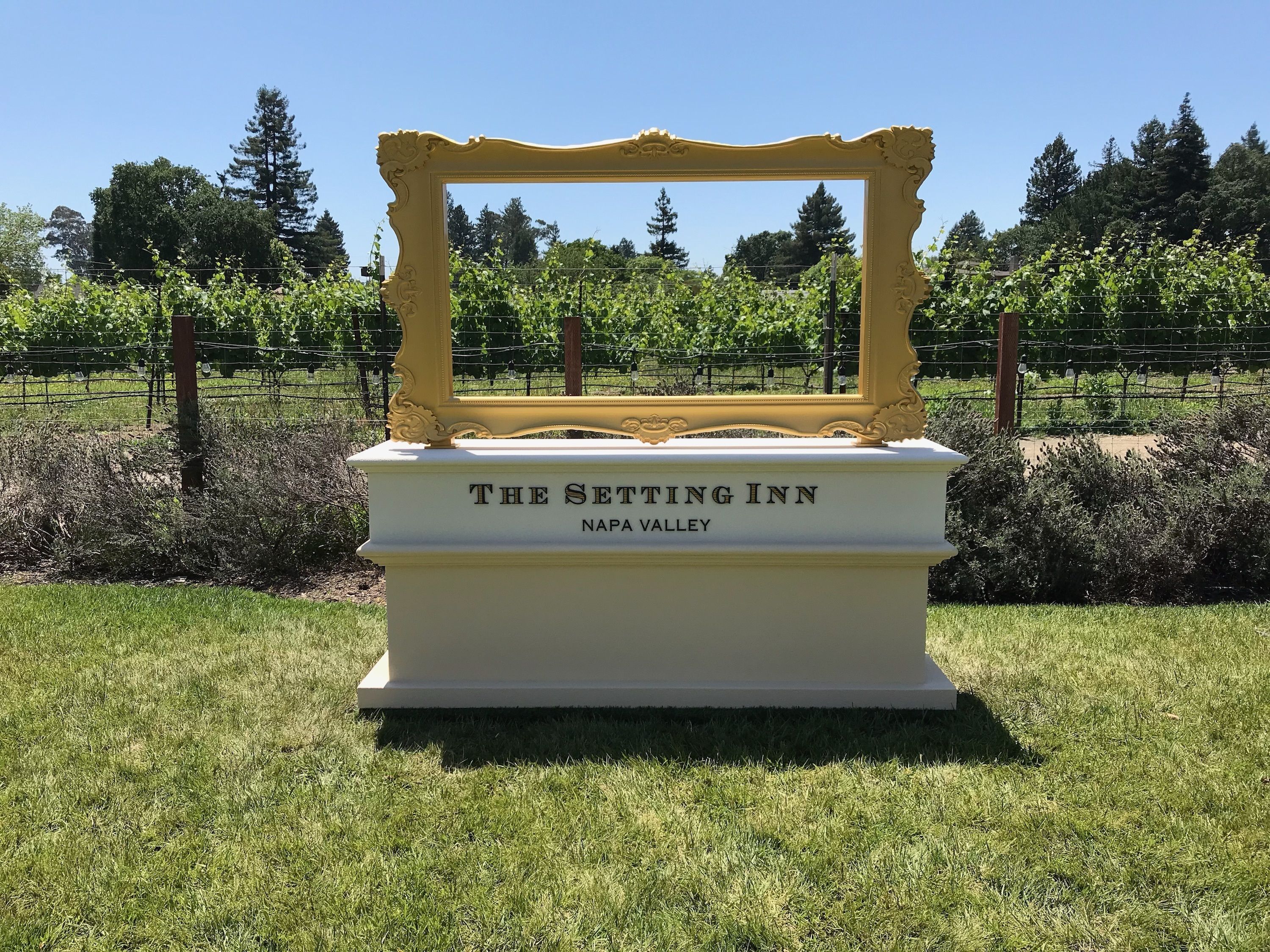 A white monument with "THE SETTING INN NAPA VALLEY" engraved on it, surrounded by grass and a vineyard in the background.