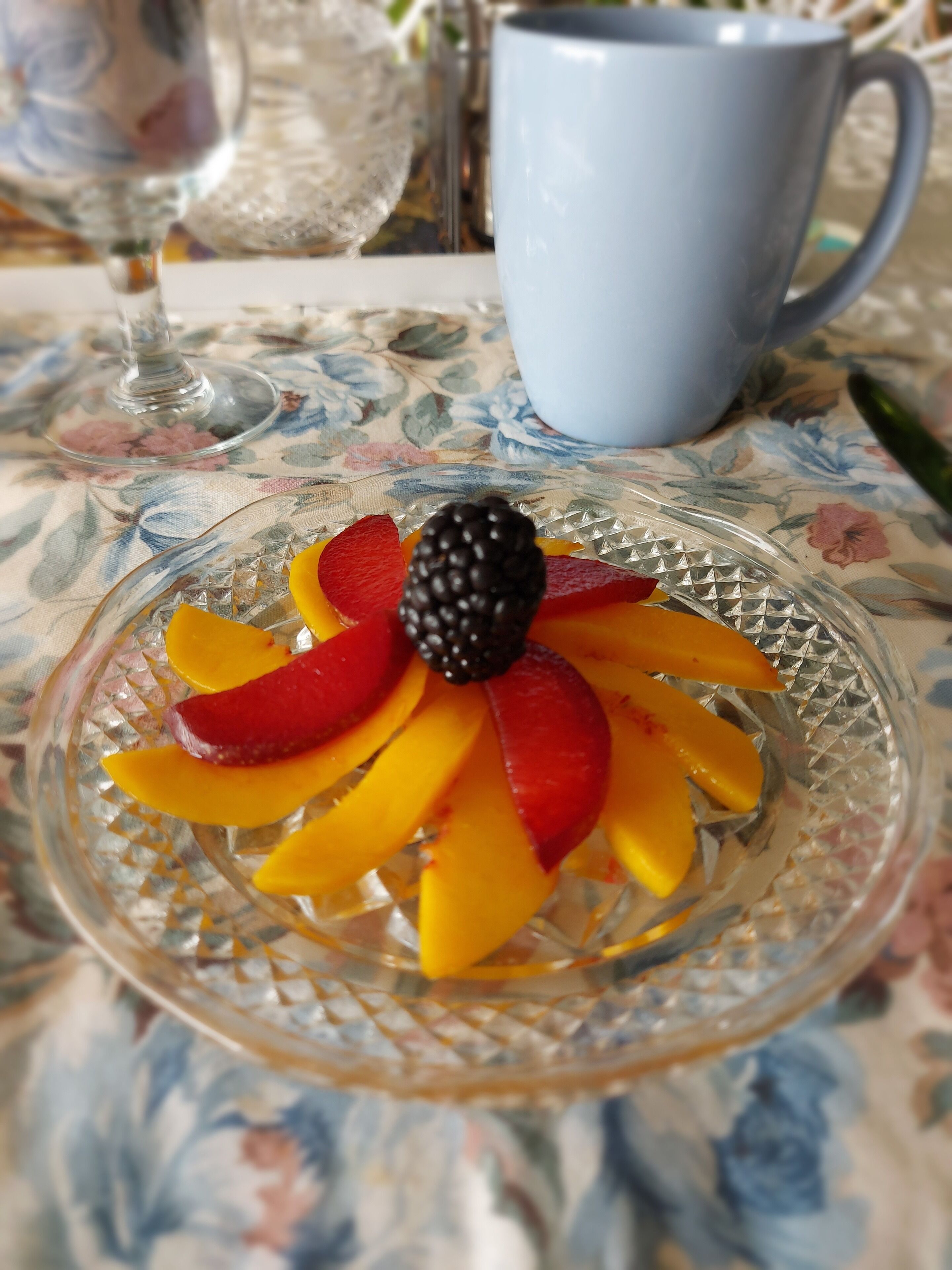 Uma tábua de vidro com frutas fatiadas e um framboeso sobre uma toalha de mesa florida.
